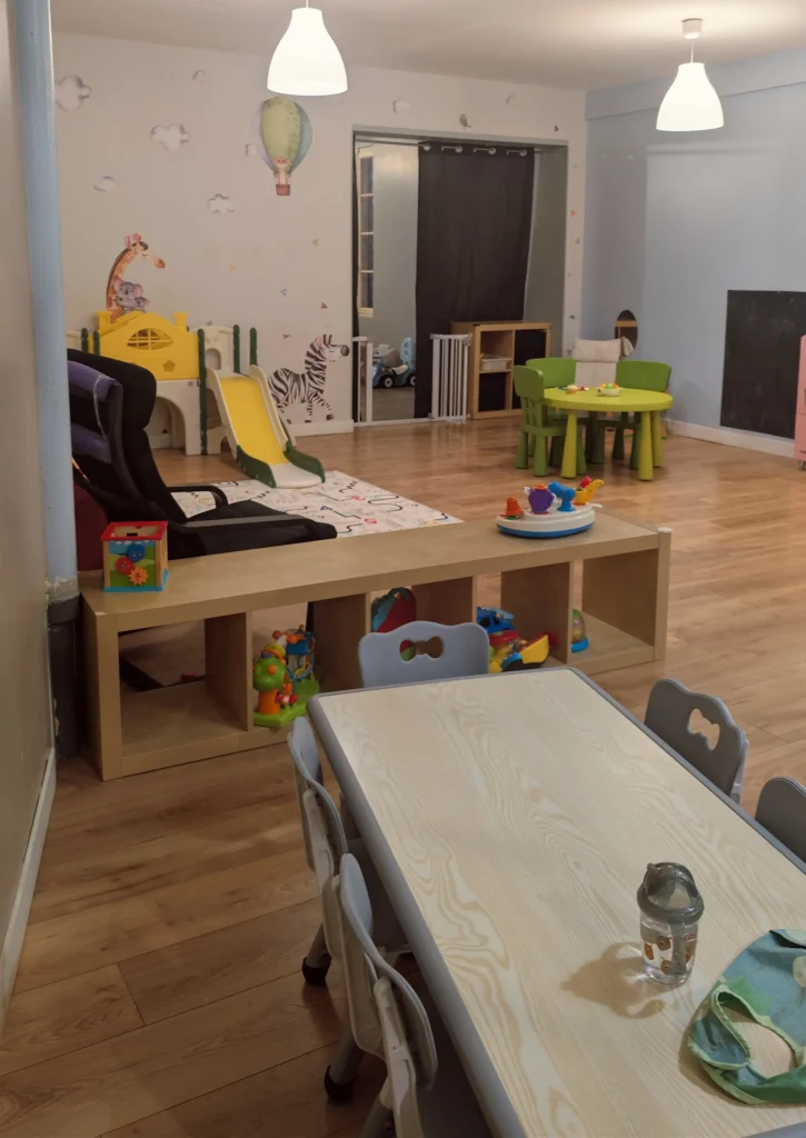 Perspective depuis le plan de travail de la cuisine donnant sur la grande pièce de vie lumineuse et organisée de la MAM Les Chérubins à Perpignan, avec les chaises hautes et les zones de jeux.